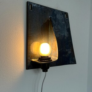 May include: A wall-mounted light fixture with a dark blue, square metal backplate. The fixture features a teardrop-shaped light holder and a round, glowing lightbulb. A black base and a gray cord complete the design.