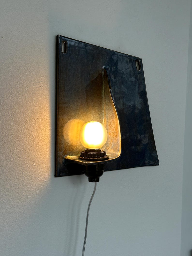 May include: A wall-mounted light fixture with a dark blue, square metal backplate. The fixture features a teardrop-shaped light holder and a round, glowing lightbulb. A black base and a gray cord complete the design.