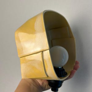 May include: A yellow ceramic wall lamp with a curved, folded design. The lamp features a smooth, glossy finish and a white spherical light bulb secured in a black socket at the base. The structure has a distinct layered appearance with visible seams.