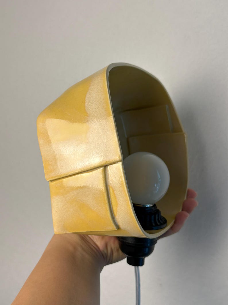 May include: A yellow ceramic wall lamp with a curved, folded design. The lamp features a smooth, glossy finish and a white spherical light bulb secured in a black socket at the base. The structure has a distinct layered appearance with visible seams.