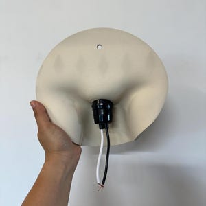 May include: A cream-colored, semi-circular wall lamp shade featuring a smooth, matte finish. The center of the shade contains a black plastic light socket with two exposed wires, one white and one black, extending downward from the base.