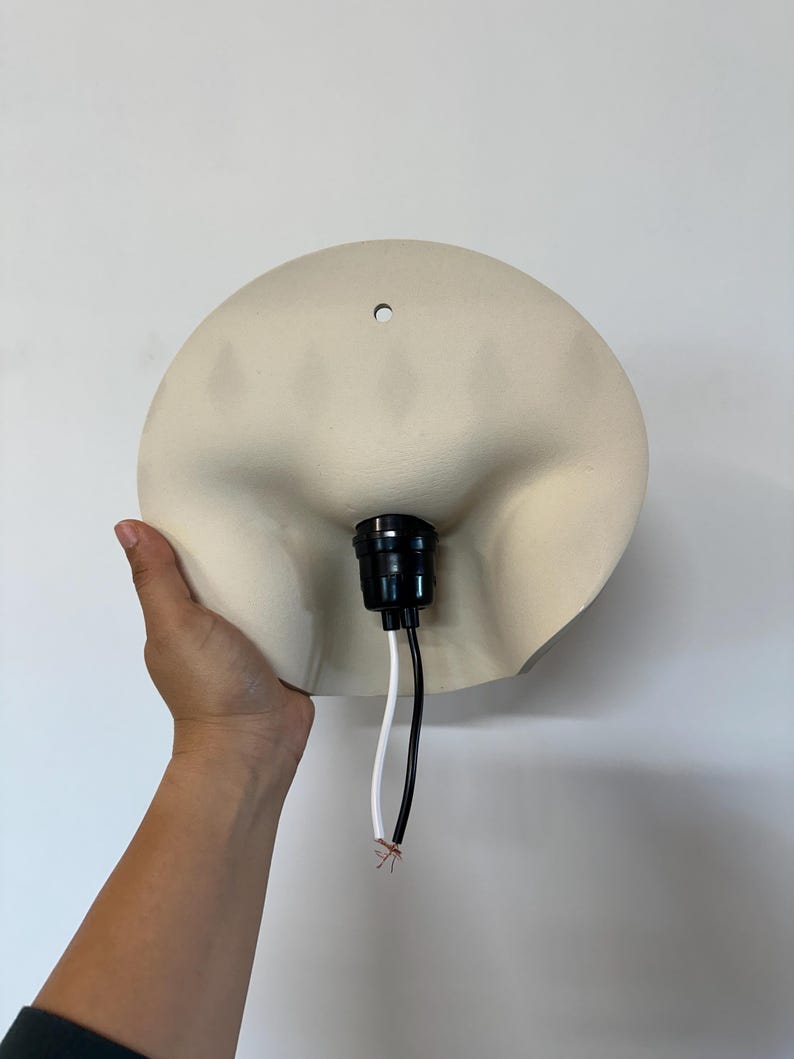 May include: A cream-colored, semi-circular wall lamp shade featuring a smooth, matte finish. The center of the shade contains a black plastic light socket with two exposed wires, one white and one black, extending downward from the base.