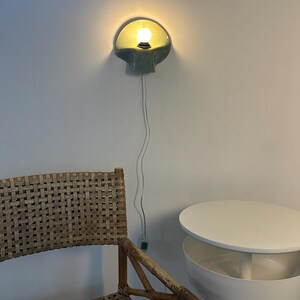 Puede incluir: Un aplique de pared de cer&aacute;mica con acabado verde salvia brillante y forma de concha, que cuenta con una bombilla encendida. Un cable el&eacute;ctrico blanco y fino cuelga verticalmente desde la l&aacute;mpara hasta un peque&ntilde;o interruptor en la pared.