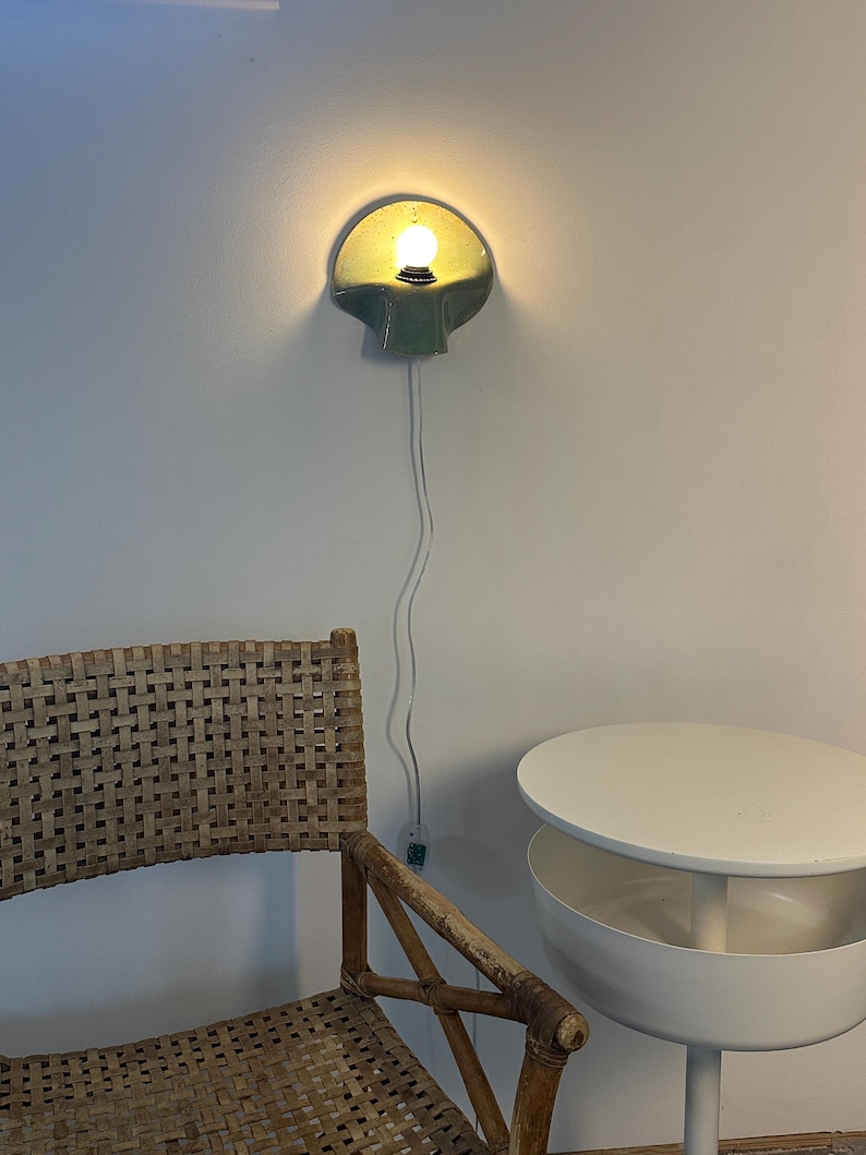Puede incluir: Un aplique de pared de cer&aacute;mica con acabado verde salvia brillante y forma de concha, que cuenta con una bombilla encendida. Un cable el&eacute;ctrico blanco y fino cuelga verticalmente desde la l&aacute;mpara hasta un peque&ntilde;o interruptor en la pared.