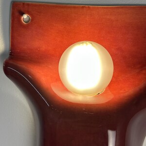 Pode incluir: Um candeeiro de parede em cer&acirc;mica com um acabamento vidrado brilhante em vermelho-alaranjado profundo e uma forma org&acirc;nica curva. Uma l&acirc;mpada redonda de vidro branco fosco est&aacute; centrada, emitindo um brilho quente e luminoso.