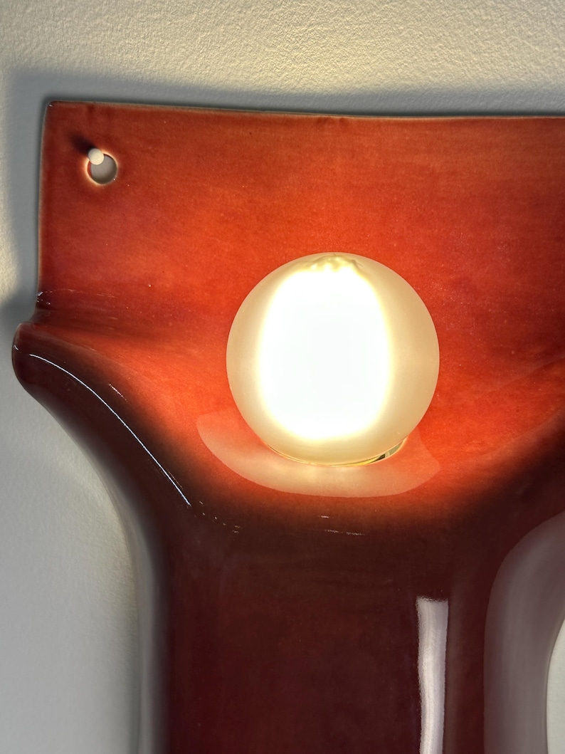 Pode incluir: Um candeeiro de parede em cer&acirc;mica com um acabamento vidrado brilhante em vermelho-alaranjado profundo e uma forma org&acirc;nica curva. Uma l&acirc;mpada redonda de vidro branco fosco est&aacute; centrada, emitindo um brilho quente e luminoso.