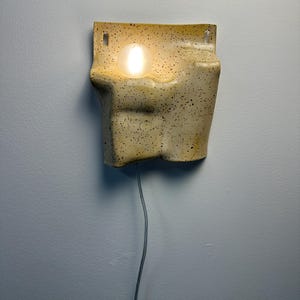 May include: A wall-mounted ceramic sconce with a speckled cream and yellow finish. The fixture features an irregular, organic shape with a recessed circular opening that holds a glowing, spherical light bulb. A thin power cord hangs from the bottom.
