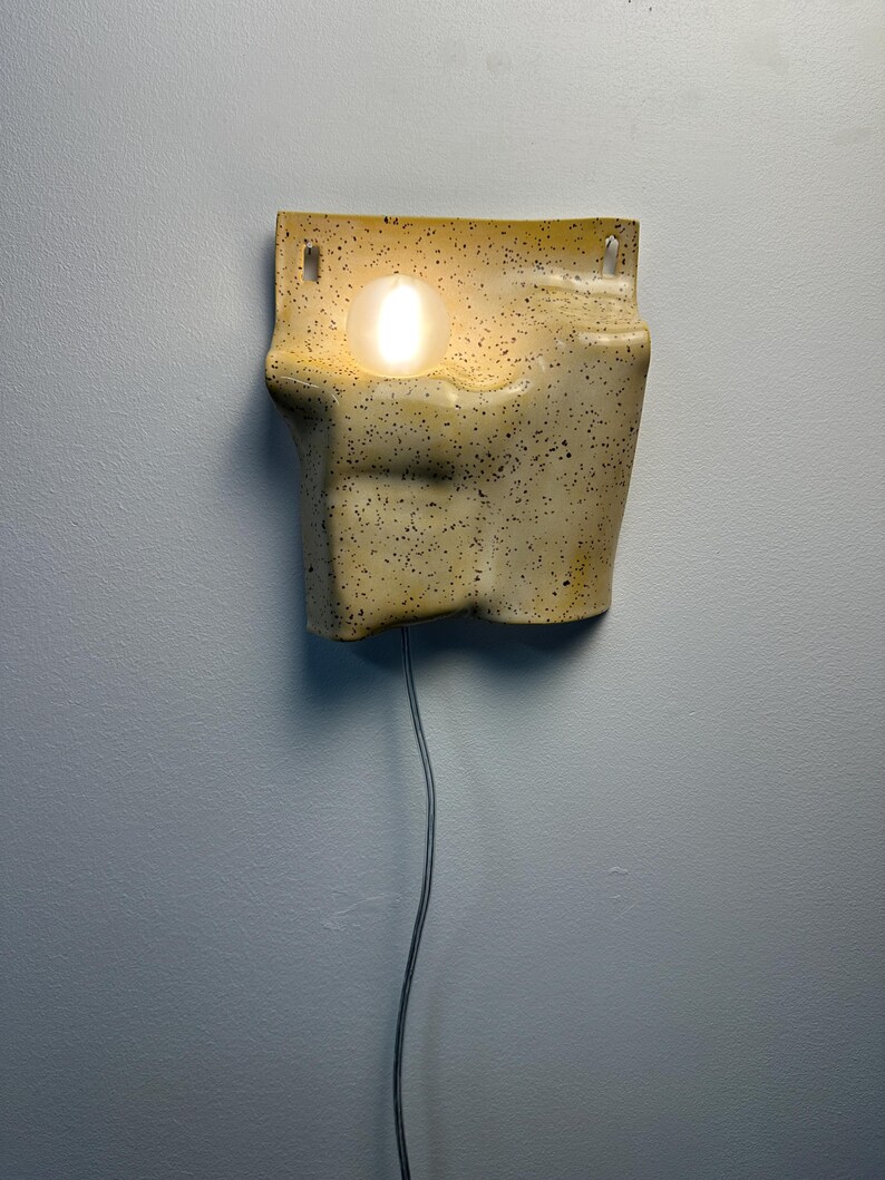 May include: A wall-mounted ceramic sconce with a speckled cream and yellow finish. The fixture features an irregular, organic shape with a recessed circular opening that holds a glowing, spherical light bulb. A thin power cord hangs from the bottom.