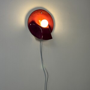 May include: A circular wall sconce featuring a glossy, deep red and burgundy gradient finish. The fixture has a curved, shell-like form with a central, glowing white spherical light bulb. A thin, transparent power cord hangs vertically downwards.