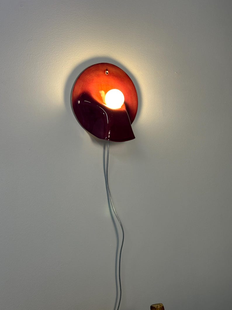 May include: A circular wall sconce featuring a glossy, deep red and burgundy gradient finish. The fixture has a curved, shell-like form with a central, glowing white spherical light bulb. A thin, transparent power cord hangs vertically downwards.