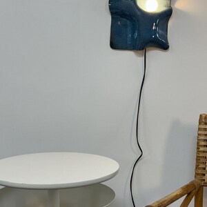 May include: A wall-mounted lamp with a glossy, irregular, dark blue ceramic shade that features a bright, glowing light bulb. The fixture is connected to a long, thin black electrical cord that hangs vertically down the wall.