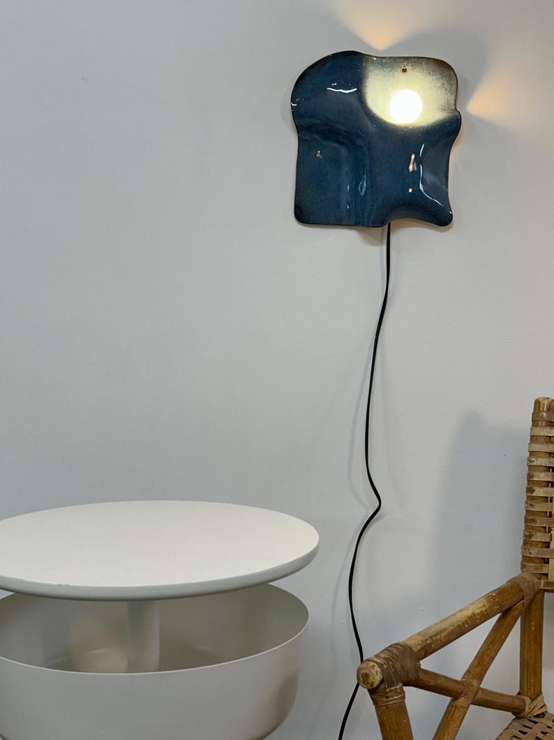 May include: A wall-mounted lamp with a glossy, irregular, dark blue ceramic shade that features a bright, glowing light bulb. The fixture is connected to a long, thin black electrical cord that hangs vertically down the wall.