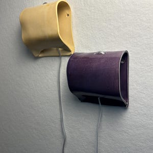 May include: Two wall-mounted lamps with curved, semi-cylindrical shades. One lamp is a matte light beige, and the other is a glossy deep purple. Each lamp features a silver metal ring at the top and a thin gray electrical cord extending downward.