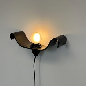 May include: A black wall sconce with a curved, wave-like design. The sconce holds a round, illuminated light bulb. A black cord extends downward from the fixture. The sconce is mounted on a light gray wall.