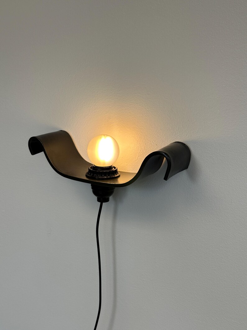 May include: A black wall sconce with a curved, wave-like design. The sconce holds a round, illuminated light bulb. A black cord extends downward from the fixture. The sconce is mounted on a light gray wall.