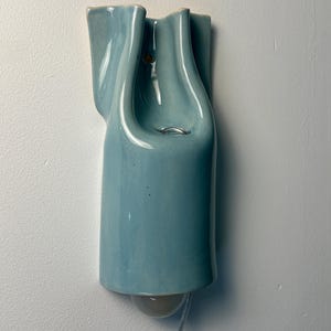 May include: A wall-mounted light fixture made of glossy, light blue ceramic with a fluid, organic shape. The piece features a central opening with a metal ring and a rounded light bulb extending from the bottom, connected by a clear power cord.