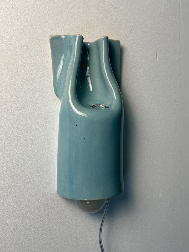 May include: A wall-mounted light fixture made of glossy, light blue ceramic with a fluid, organic shape. The piece features a central opening with a metal ring and a rounded light bulb extending from the bottom, connected by a clear power cord.