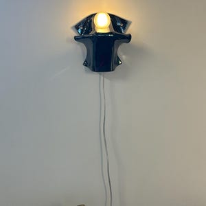 May include: A dark blue ceramic wall sconce with a glossy finish and a curved, organic shape. The fixture holds a single glowing round light bulb at the top center. A thin, transparent electrical cord hangs vertically downward from the base.