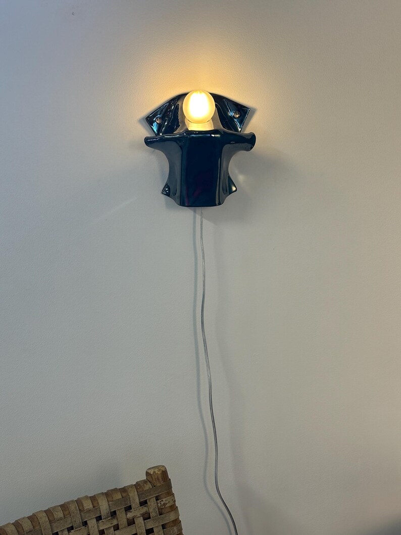 May include: A dark blue ceramic wall sconce with a glossy finish and a curved, organic shape. The fixture holds a single glowing round light bulb at the top center. A thin, transparent electrical cord hangs vertically downward from the base.
