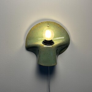 Puede incluir: Un aplique de pared con una pantalla de cer&aacute;mica curva en forma de concha de color verde claro con motas oscuras. Un casquillo negro central sostiene una bombilla redonda esmerilada encendida. Un cable blanco fino cuelga desde abajo.
