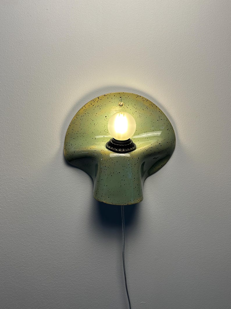 Puede incluir: Un aplique de pared con una pantalla de cer&aacute;mica curva en forma de concha de color verde claro con motas oscuras. Un casquillo negro central sostiene una bombilla redonda esmerilada encendida. Un cable blanco fino cuelga desde abajo.