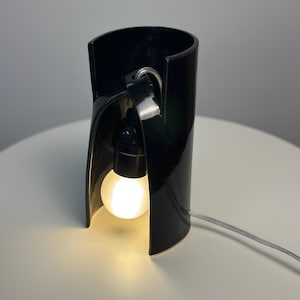 May include: A glossy black ceramic table lamp with a cylindrical, open-front design. A black socket holds a glowing, round white light bulb suspended inside the curved structure, connected to a thin, clear electrical cord extending outward.