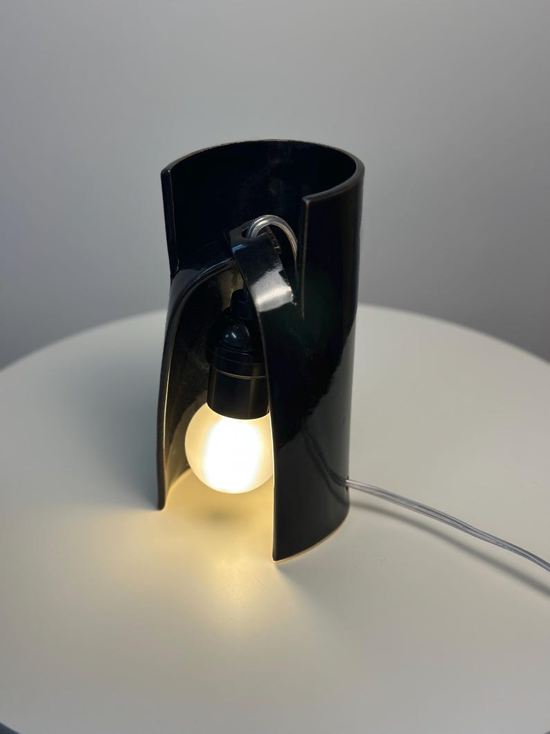May include: A glossy black ceramic table lamp with a cylindrical, open-front design. A black socket holds a glowing, round white light bulb suspended inside the curved structure, connected to a thin, clear electrical cord extending outward.