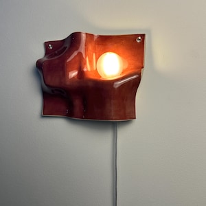 May include: A wall-mounted sconce featuring a glossy, deep reddish-brown ceramic shade with an organic, undulating wave-like form. The fixture includes a single glowing spherical light bulb nestled within a recessed curve and a thin power cord.
