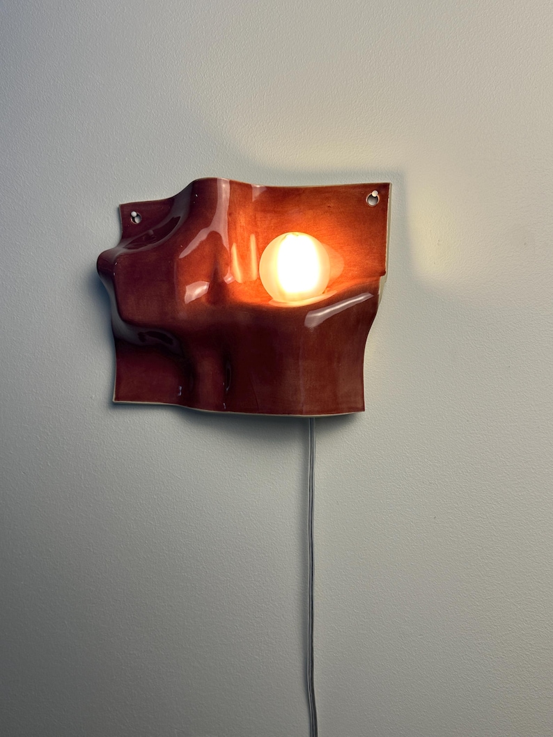 May include: A wall-mounted sconce featuring a glossy, deep reddish-brown ceramic shade with an organic, undulating wave-like form. The fixture includes a single glowing spherical light bulb nestled within a recessed curve and a thin power cord.