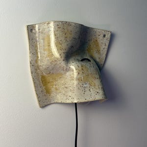May include: A wall-mounted ceramic sconce with a curved, irregular shape. The surface features a speckled cream glaze with yellow-toned brushstrokes. A black electrical cord extends from the bottom, and two small holes are visible at the top corners.
