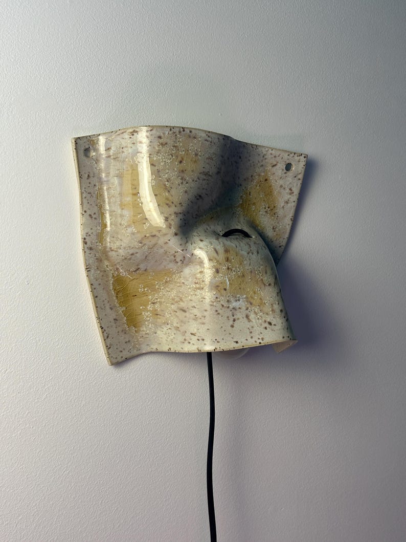 May include: A wall-mounted ceramic sconce with a curved, irregular shape. The surface features a speckled cream glaze with yellow-toned brushstrokes. A black electrical cord extends from the bottom, and two small holes are visible at the top corners.