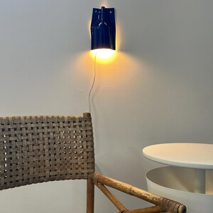 Puede incluir: Un aplique de pared de color azul oscuro con una pantalla cil&iacute;ndrica que emite una luz amarilla c&aacute;lida. La l&aacute;mpara est&aacute; fijada a la pared mediante una placa trasera rectangular plana y tiene un cable de alimentaci&oacute;n blanco que baja.