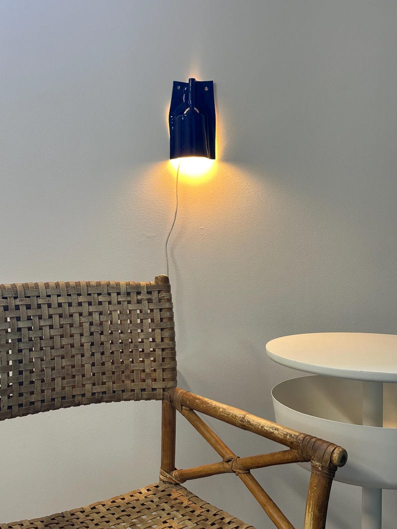 Puede incluir: Un aplique de pared de color azul oscuro con una pantalla cil&iacute;ndrica que emite una luz amarilla c&aacute;lida. La l&aacute;mpara est&aacute; fijada a la pared mediante una placa trasera rectangular plana y tiene un cable de alimentaci&oacute;n blanco que baja.