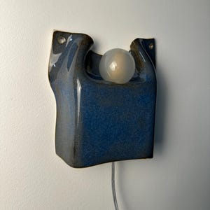 May include: A wall-mounted ceramic light fixture with a glossy, deep blue glaze. The sculptural form features an organic, curved shape with two upper mounting holes and a central circular opening holding a single, frosted white spherical light bulb.