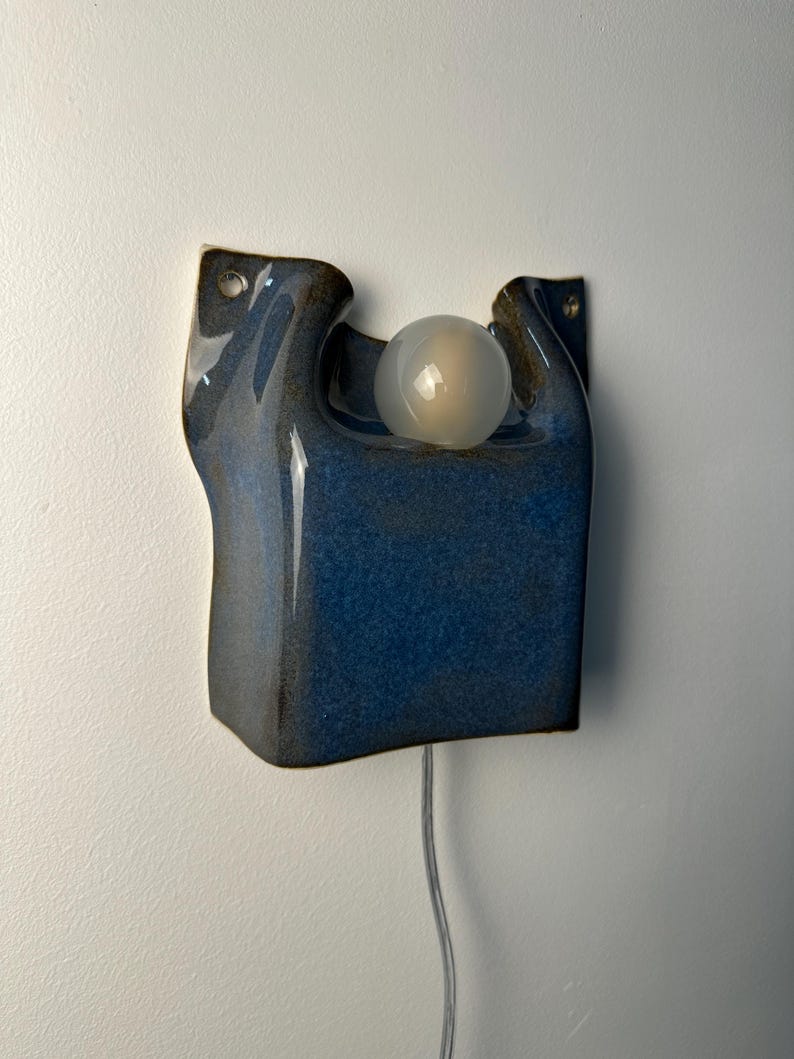 May include: A wall-mounted ceramic light fixture with a glossy, deep blue glaze. The sculptural form features an organic, curved shape with two upper mounting holes and a central circular opening holding a single, frosted white spherical light bulb.