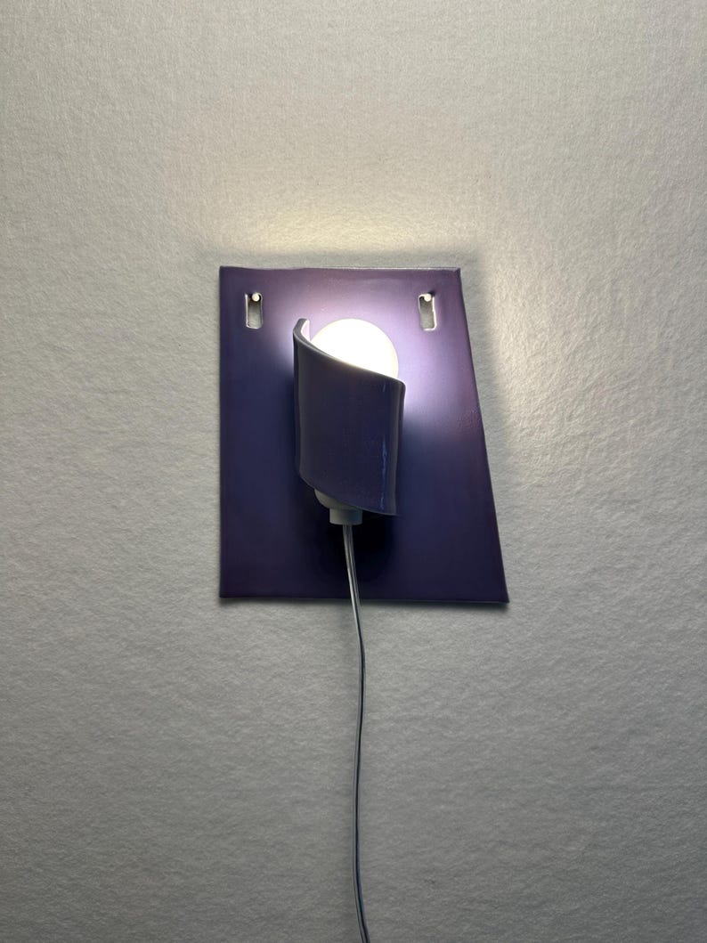 Pode incluir: Uma arandela de parede moderna com uma placa traseira retangular roxa escura. Uma sombra curva, roxa escura, abriga uma l&acirc;mpada redonda, branca e brilhante. A arandela &eacute; conectada com um fio cinza fino.