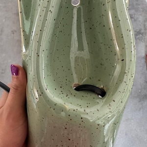 May include: A sage green ceramic wall sconce with a speckled finish and a glossy glaze. The fixture features a curved, elongated body with a circular mounting hole at the top and a central opening at the bottom for a black electrical cord to pass through.