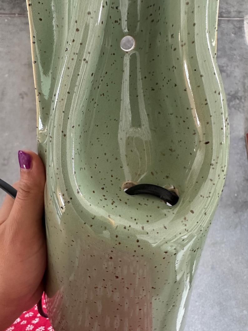 May include: A sage green ceramic wall sconce with a speckled finish and a glossy glaze. The fixture features a curved, elongated body with a circular mounting hole at the top and a central opening at the bottom for a black electrical cord to pass through.