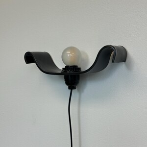 May include: A black, wall-mounted light fixture with a curved, wave-like design. The fixture holds a round, frosted light bulb. A black cord extends downward from the fixture. The light is mounted on a white wall.