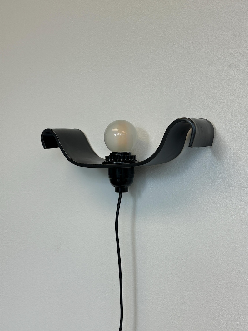 May include: A black, wall-mounted light fixture with a curved, wave-like design. The fixture holds a round, frosted light bulb. A black cord extends downward from the fixture. The light is mounted on a white wall.
