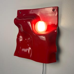 May include: A glossy red wall-mounted lamp featuring a square, undulating, wave-like surface with a central circular cutout. A single glowing white spherical light bulb is recessed within the opening, and a thin power cord extends from the bottom.