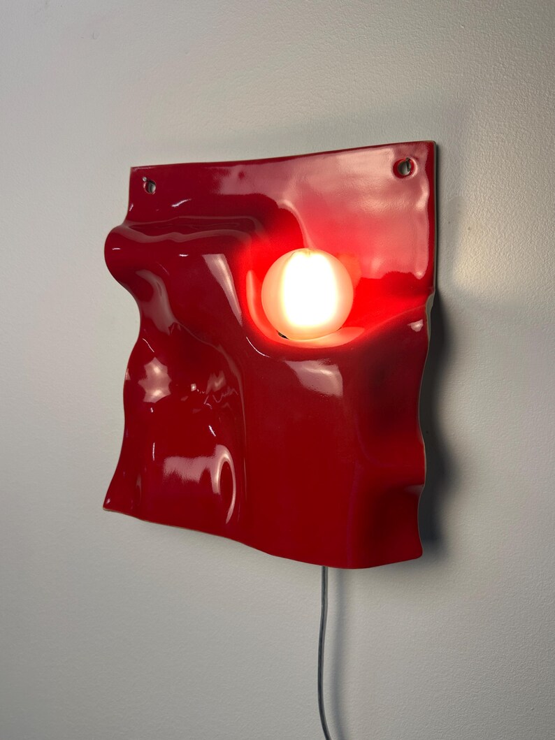 May include: A glossy red wall-mounted lamp featuring a square, undulating, wave-like surface with a central circular cutout. A single glowing white spherical light bulb is recessed within the opening, and a thin power cord extends from the bottom.
