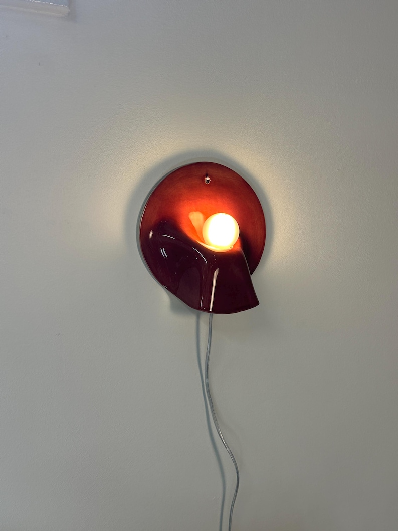 May include: A circular wall lamp with a deep red, glossy finish and a curved, sculptural form. A single, warm-toned spherical light bulb is positioned within the centre of the fixture, emitting a soft glow. A thin, light-coloured power cord hangs down.
