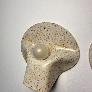 May include: A wall-mounted light fitting featuring a curved, speckled cream-coloured ceramic shade with brown flecks and a central circular glass bulb. The ceramic shade has a wide, flared shape with a small hole at the top for mounting purposes.