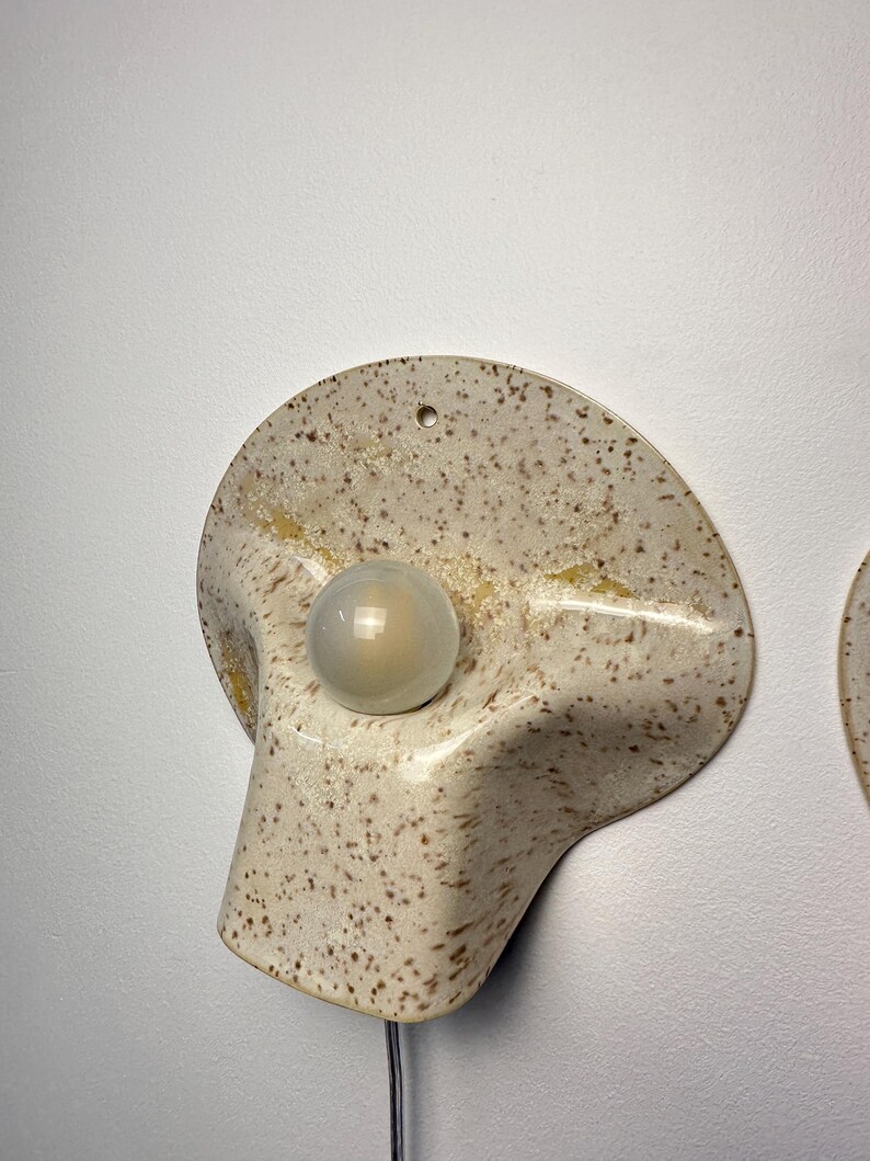 May include: A wall-mounted light fitting featuring a curved, speckled cream-coloured ceramic shade with brown flecks and a central circular glass bulb. The ceramic shade has a wide, flared shape with a small hole at the top for mounting purposes.