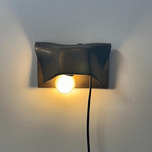 Pode incluir: Um candeeiro de parede com um abajur de metal escuro e curvo sobre uma placa retangular. Uma l&acirc;mpada redonda e iluminada est&aacute; posicionada sob o abajur, e um cabo de alimenta&ccedil;&atilde;o preto pende verticalmente da lumin&aacute;ria ao longo da parede.