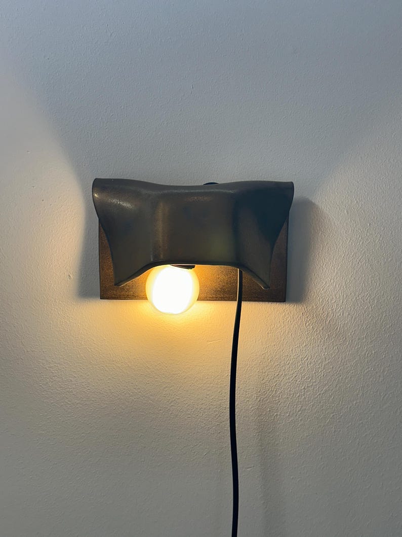 Pode incluir: Um candeeiro de parede com um abajur de metal escuro e curvo sobre uma placa retangular. Uma l&acirc;mpada redonda e iluminada est&aacute; posicionada sob o abajur, e um cabo de alimenta&ccedil;&atilde;o preto pende verticalmente da lumin&aacute;ria ao longo da parede.