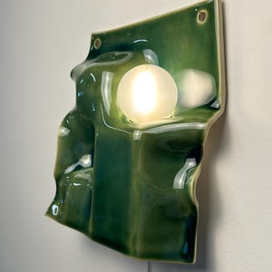 May include: A wall-mounted ceramic light fixture with a glossy, deep green glazed finish. The surface features an irregular, undulating, and folded form. A single, round, frosted white glass bulb is embedded within the ceramic structure.