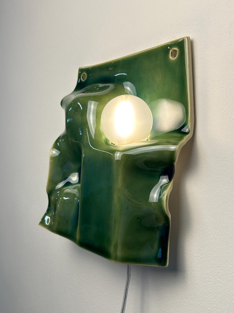May include: A wall-mounted ceramic light fixture with a glossy, deep green glazed finish. The surface features an irregular, undulating, and folded form. A single, round, frosted white glass bulb is embedded within the ceramic structure.