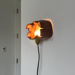 May include: A wall-mounted, brown, decorative light fixture with an orange glow. The fixture has an abstract, organic shape and a visible light bulb. A black cord extends downward from the fixture.